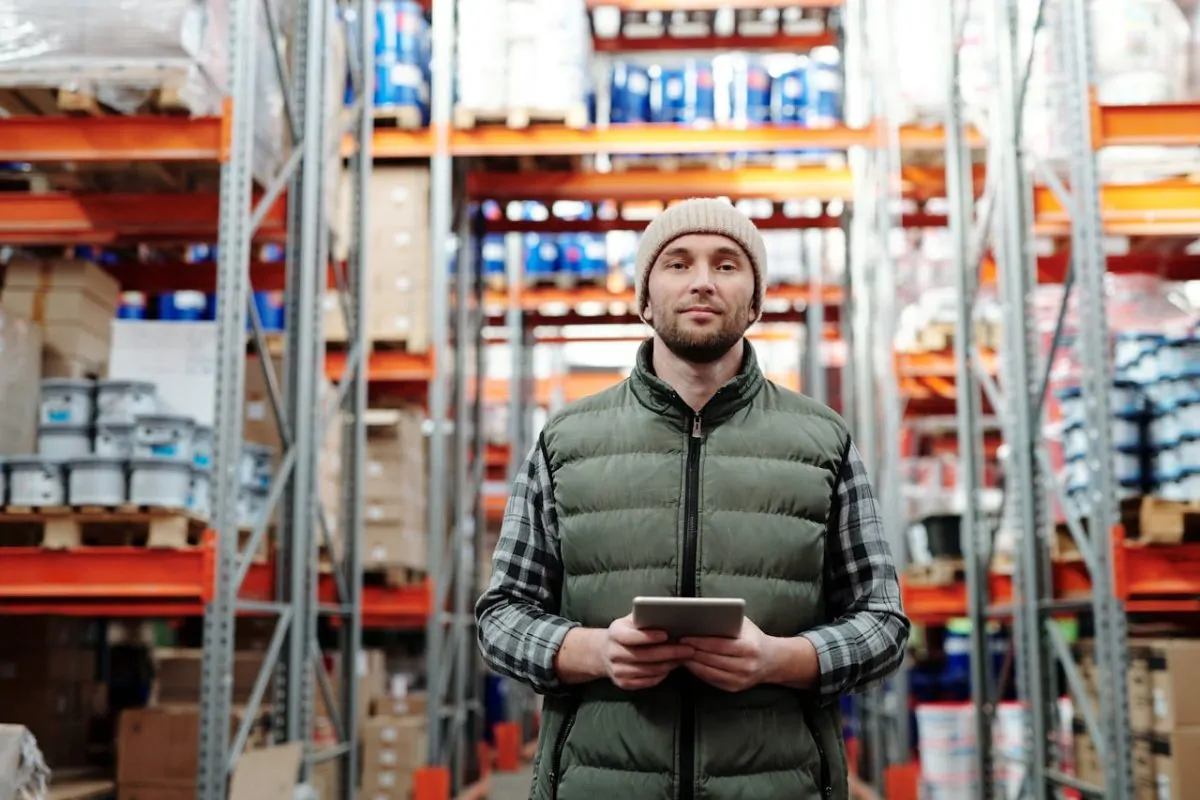 Warehouse manager holding a tablet, standing in a well-stocked storage facility with shelves filled with inventory and industrial supplies.
